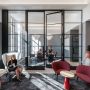 The alumni lounge is shown with a tall, glass, doored wall separating a seating area with four chairs from a conference area with a table and chairs. In the foreground, a woman sits in a chair, reading. Three other people populate the scene.
