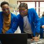 Two young men in blue coats and goggles look at a laptop together. A woman is in the background