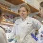 Nicole De Nisco and two others chat at a biology lab bench. All three are wearing white lab coats and hair in ponytails.