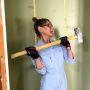 Jessica Banks wields a sledgehammer in position to strike. She is wearing gloves and coveralls and looking toward a wall with large holes in it.