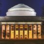 MIT's Building 10 is shown lit up at night. The dome of the building looks almost white against the dark blue sky. Below, a row of columns front the building's portico, and a glow of yellow light shines through windows from the interior.  Above the columns are the words "Massachusetts Institute of Technology."