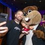 A man in a black T-shirt takes a selfie with Tim the Beaver. Some people are visible in the dark background.
