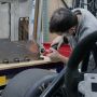 A man leans over a workbench holding a grinder against a metal surface. Sparks fly. A tire and a steering wheel are visible in the foreground.