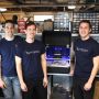 Three men in navy T-shirts (two shirts clearly read Optigon) pose in a workshop. On the workbench between two of them is an open black case. Parts inside appear blue and gray.