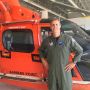 Gene McGuinness stands in front of a red helicopter wearing an olive flightsuit with a Coast Guard patch on it.