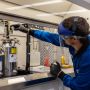Zach Goodman, wearing full lab protection, adjusts a gauge above a stainless steel cylinder in a lab.