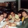 Five men sit around a table eating pizza with forks. There are some cups on the table and a suitcase and some boxes are visible in the background.