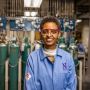 Sossina Haile portrait. She is wearing a blue lab coat featuring a purple N. She is standing in front of an array of narrow green tanks and other equipment.