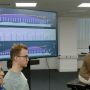 A large screen showing a variety of charts is at left in a room where Elaine Chew is playing piano and two students, at left, listen.