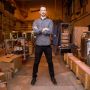 Zachary Hartwig stands arms crossed in what looks like a large workshop. Nitrogen tanks, boxes, ladders, wires, and other materials can be seen in the background.