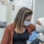 A mother hugs her young daughter while she receives cancer treatment. Both are masked, and the child holds a plush toy