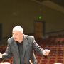 Jonathan Pasternack is shown conducting. He holds a baton and the seats of a theater can be seen in the background.