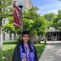 Tara Venkatadri appears in her graduation cap and gown in front of McCormick Hall at MIT. A red banner with the dorm name is attached to a light pole behind her.