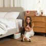 Loewen Cavill sits on the floor next to a bed holding a small device. There is a larger device on the floor next to her and a pad on the bed. Behind Cavill, a cardboard box rests on a chest of drawers.