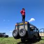 Jamila Smith-Dell Stands on the top of a Land Rover on a safari, looking off to the right with blue sky and grass below