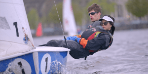 Two MIT students lean out of a small sailboat with their bodies half hanging over the Charles River