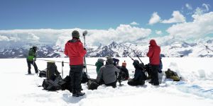 A photo of a crew of people sitting and standing in snow with filming equipment and with mountains in the background