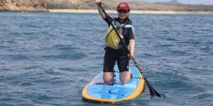 Woman wearing a wetsuit kneeling on a paddleboard in the ocean with a beach and rocky coastline behind her