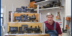 Ed Hume, dressed in overalls, sits next to a three-level display of model trains in his workshop, with another displayed on a shelf on the wall behind him.