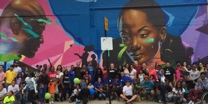 A large group on a sidewalk in front of a mural of a man and woman's faces