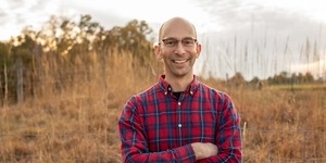 Smiling man with glasses, no hair, and plaid shirt standing in a field with arms crossed 