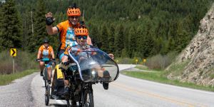 Photo of Steve Zuckerman (waving) and Deb Meyerson riding a tandem bike on a road with trees in the background and another biker behind them