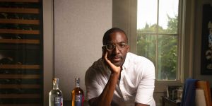 Photo of Marc-Kwesi Farrell leaning on a counter with two bottles of rum to the left a window behind him on the right and a wine fridge on the left