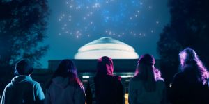 Four people with their backs facing the camera looking at the MIT dome on Killian Court at night with lights in the sky 