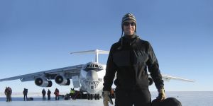 Greg Maud is shown in foreground wearing winter hat. Airplane is in background with a few people beside it. Clear sky. Snowy ground.