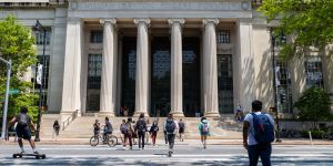 A photo of 77 Mass Ave building at MIT, buildings with columns and students crossing the street in front of the building