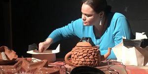 Erin Genia is shown from chest up leaning over an array of clay artwork, including a teapot and a large star.