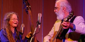 Sharon Horovitch, left, and Jim Muller are shown from the waist up with microphones in foreground. Horovitch is holding the neck of a bass fiddle. Muller is playing a guitar. They are looking at each other.