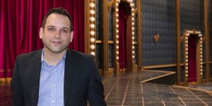 Daniel Perez stands in a theater . The stage, a red curtain, and two doorways edged with glowing lightbulbs are in the background.