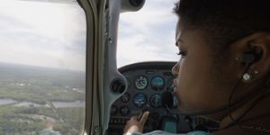 A close profile of Elissa Gibson with the controls of a plane in front of her and an aerial view of the landscape visible below