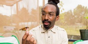 Andre Green observes a monarch butterfly as it sits on a small tube he holds between his thumb and finger. Two mesh containers can be seen to his left and right. 