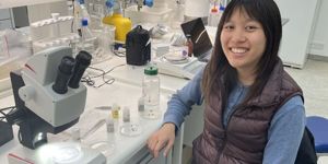 Andrea Lo sits in a lab with a microscope and other lab equipment visible on the counter.