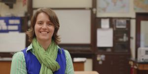Shannon Morey portrait shows her in a classroom, which is blurry in the background. She wears a green scarf, blue vest, and striped green collared shirt.