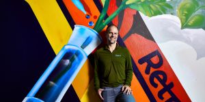 Joe Betts-LaCroix leans against a wall with a mural on it, featuring a test tube with a green stem coming out the top and yellow orange and red lines, with the word Retro written on the red line