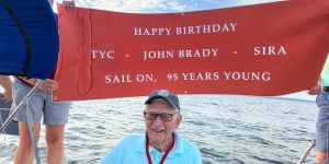 John Brady wears a blue shirt and a baseball cap. A huge red banner held up behind him reads, "Happy birthday - TYC - John Brady - SIRA/ Sail on, 96 Years Young." Water is in the background.