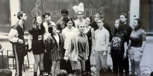 An old black and white photo shows a group of singers in front of a blackboard on which the word Chorallaries is written. The singers are wearing an assortment of garments, including a feather headress, a bandana, and a bowtie. The floor is littered with trash. The heads of some audience members appear in the foreground.