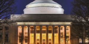 MIT's Building 10 is shown lit up at night. The dome of the building looks almost white against the dark blue sky. Below, a row of columns front the building's portico, and a glow of yellow light shines through windows from the interior.  Above the columns are the words "Massachusetts Institute of Technology."