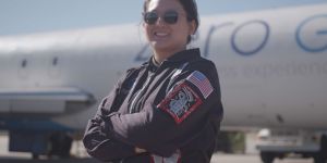 A woman in a flight suit and sunglasses stands with her arms crossed. Patches on one sleeve show an American flag and a rocket ship. A plane is visible in the background.