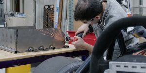 A man leans over a workbench holding a grinder against a metal surface. Sparks fly. A tire and a steering wheel are visible in the foreground.