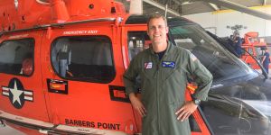 Gene McGuinness stands in front of a red helicopter wearing an olive flightsuit with a Coast Guard patch on it.