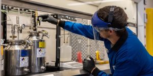 Zach Goodman, wearing full lab protection, adjusts a gauge above a stainless steel cylinder in a lab.