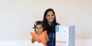 A photo of Amrita Saigal sitting on a white fuzzy rug with boxes of kudos diapers in front of her and a child sitting on one of the boxes