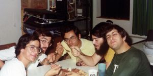 Five men sit around a table eating pizza with forks. There are some cups on the table and a suitcase and some boxes are visible in the background.