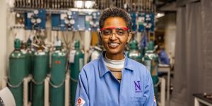 Sossina Haile portrait. She is wearing a blue lab coat featuring a purple N. She is standing in front of an array of narrow green tanks and other equipment.