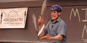 A photo of David Rush in a classroom with a blackboard behind him wearing a helmet and juggling knives