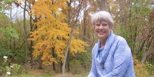 Sara Clope Long is shown facing the camera. She is outside and resting her wrist on a black railing. The area in the background is wooded.